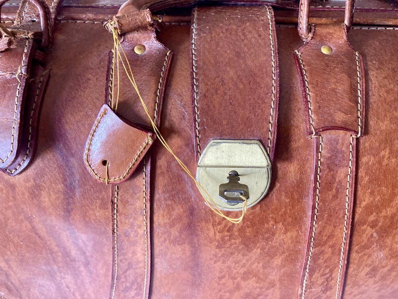 Puede incluir: Primer plano de un bolso de cuero marr&oacute;n vintage con un cierre de metal y correas de cuero. El bolso tiene un aspecto desgastado y texturizado con costuras visibles y una peque&ntilde;a etiqueta de cuero sujeta con una cuerda.