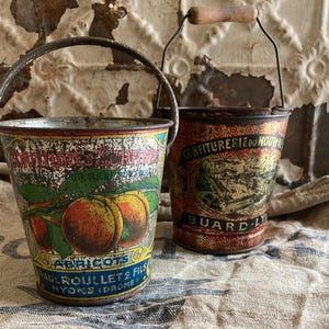May include: Two vintage metal buckets with wire handles and wooden grips. The front bucket features a colorful illustration of apricots with the text "CONFITURES SURFINES ABRICOTS PAUL ROULLET & FILS NYONS (DROME)". The back bucket has a dark red pattern.
