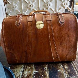 Puede incluir: Una maleta de cuero marr&oacute;n vintage con asa, cierre de lat&oacute;n y detalles cosidos. La maleta rectangular est&aacute; asegurada con correas y hebillas. El cuero tiene un aspecto texturizado. Un art&iacute;culo de viaje cl&aacute;sico.