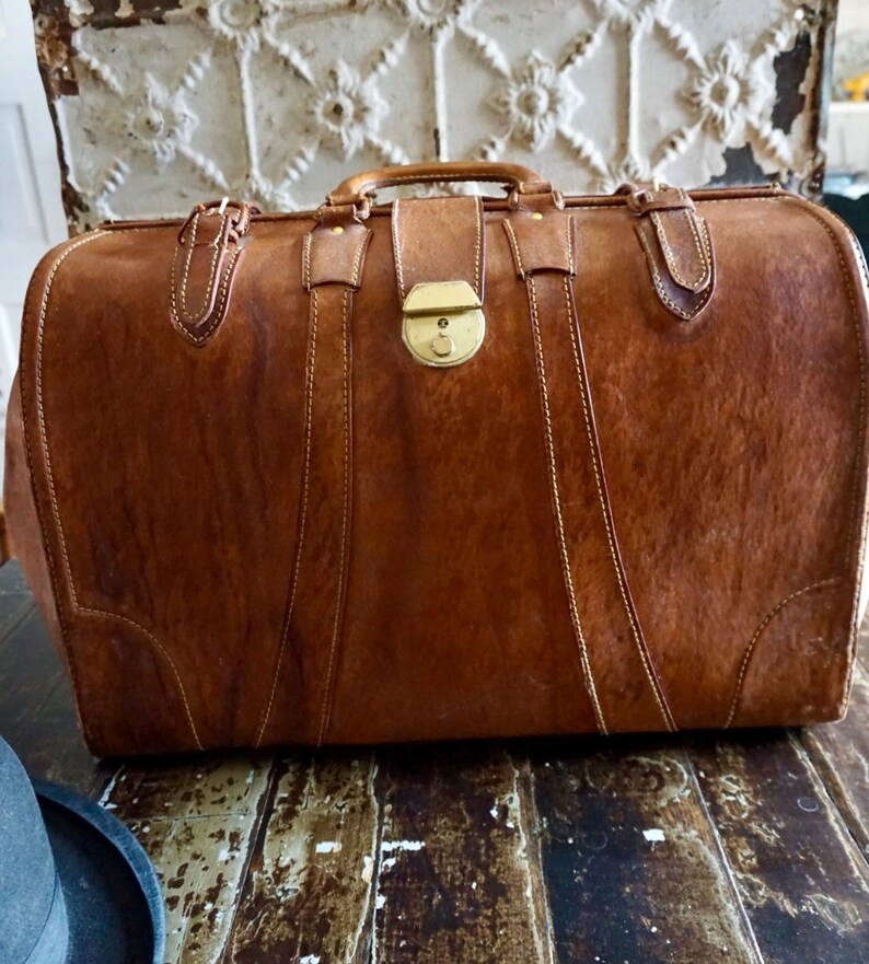 Puede incluir: Una maleta de cuero marr&oacute;n vintage con asa, cierre de lat&oacute;n y detalles cosidos. La maleta rectangular est&aacute; asegurada con correas y hebillas. El cuero tiene un aspecto texturizado. Un art&iacute;culo de viaje cl&aacute;sico.