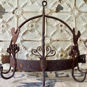 May include: A dark brown, rusted iron wall-mounted pot rack featuring a semi-circular frame with a central twisted metal support, decorative scrollwork, and two rooster silhouettes on either side, equipped with three downward-curving hooks.