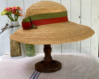 May include: A straw sun hat with a wide brim, displayed on a wooden stand. The hat features a green and red ribbon band. The hat is a light tan color and appears to be made of woven straw.