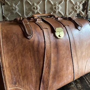 Puede incluir: Una maleta vintage de cuero marr&oacute;n con herrajes de lat&oacute;n. La maleta tiene un asa y correas con hebillas. El cuero muestra signos de desgaste, d&aacute;ndole un aspecto r&uacute;stico. La maleta se cierra con un cierre de lat&oacute;n.