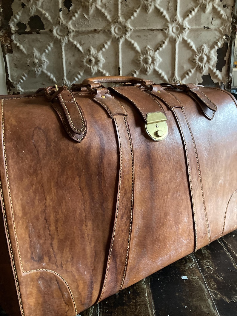 Puede incluir: Una maleta vintage de cuero marr&oacute;n con herrajes de lat&oacute;n. La maleta tiene un asa y correas con hebillas. El cuero muestra signos de desgaste, d&aacute;ndole un aspecto r&uacute;stico. La maleta se cierra con un cierre de lat&oacute;n.