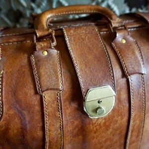 Puede incluir: Primer plano de una bolsa de viaje de cuero marr&oacute;n con asa y correas. La bolsa presenta un cierre de lat&oacute;n y detalles de costura. El cuero tiene un aspecto vintage desgastado.