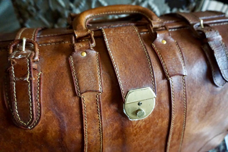 Puede incluir: Primer plano de una bolsa de viaje de cuero marr&oacute;n con asa y correas. La bolsa presenta un cierre de lat&oacute;n y detalles de costura. El cuero tiene un aspecto vintage desgastado.