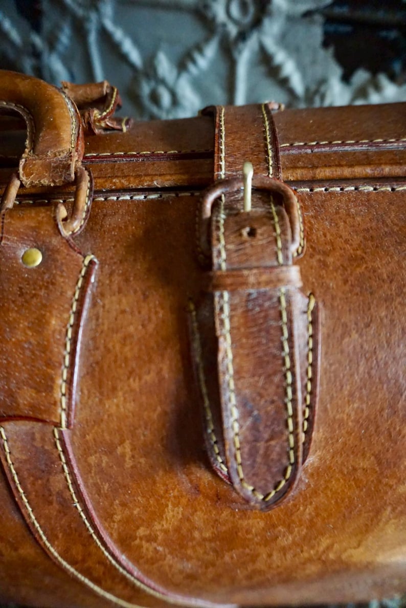 Puede incluir: Primer plano de una maleta de cuero marr&oacute;n vintage con cierre de hebilla. La maleta presenta costuras visibles en hilo amarillo en contraste, y se ve un asa en la parte superior. El cuero muestra signos de desgaste.