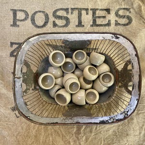 Peut inclure: Un panier ovale en fil métallique avec un bord peint en blanc vieilli, rempli de petites tasses en céramique blanc cassé. Le panier repose sur un tissu beige avec le mot "POSTES" imprimé en grandes lettres sombres.