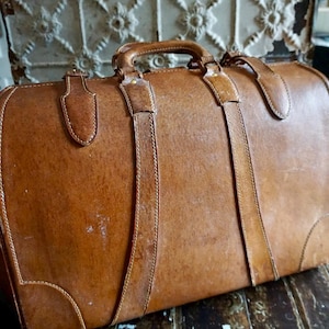 Puede incluir: Una maleta vintage de cuero marr&oacute;n con asa superior y dos correas. La maleta tiene costuras visibles y herrajes met&aacute;licos. El cuero muestra signos de desgaste, lo que aumenta su encanto antiguo. La maleta probablemente se utiliza para viajar.
