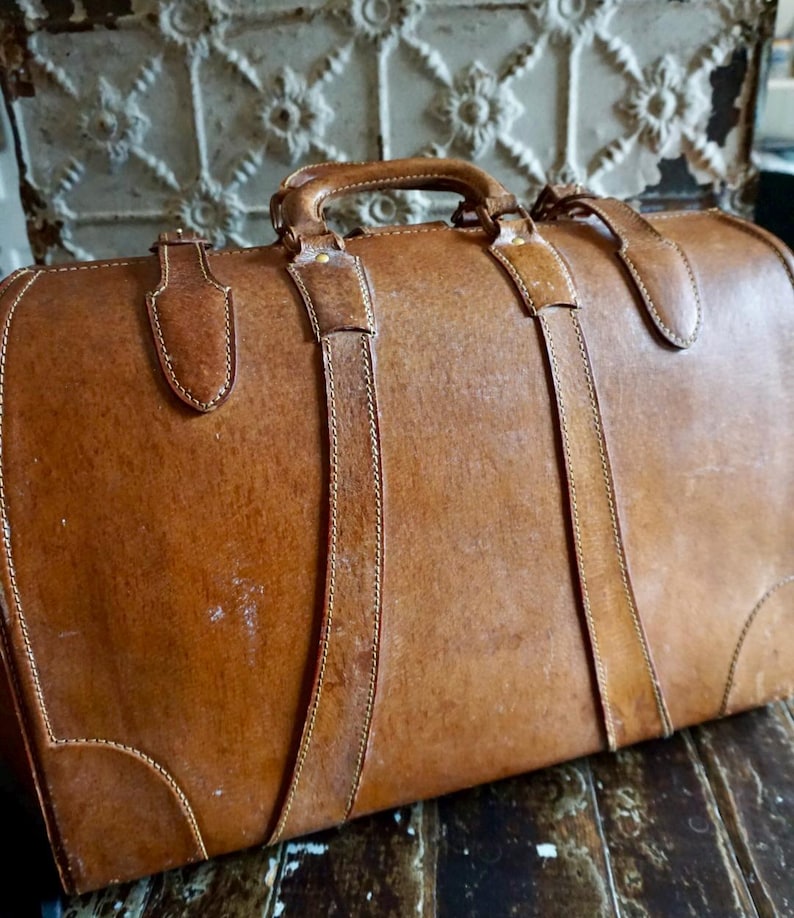 Puede incluir: Una maleta vintage de cuero marr&oacute;n con asa superior y dos correas. La maleta tiene costuras visibles y herrajes met&aacute;licos. El cuero muestra signos de desgaste, lo que aumenta su encanto antiguo. La maleta probablemente se utiliza para viajar.