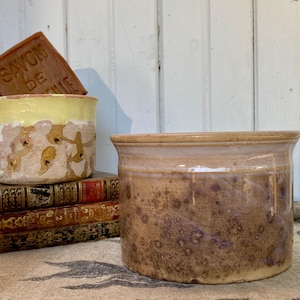 May include: A collection of ceramic containers in various sizes and designs. One container is a light brown with a purple speckled pattern. Another is a yellow and beige container with a floral design. A bar of soap with text is visible.