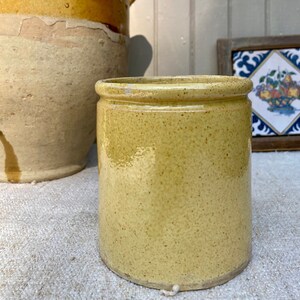 May include: A cylindrical, yellow-glazed ceramic jar with a speckled surface. The jar has a slightly flared rim and a rounded base. Another, larger, similar jar is partially visible in the background. The style suggests a vintage or antique kitchenware item.