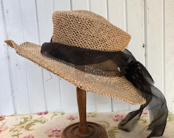 May include: A straw hat with a wide brim and a black ribbon. The hat is displayed on a wooden hat stand. The hat has a woven texture and a slightly distressed appearance, suggesting a vintage style.