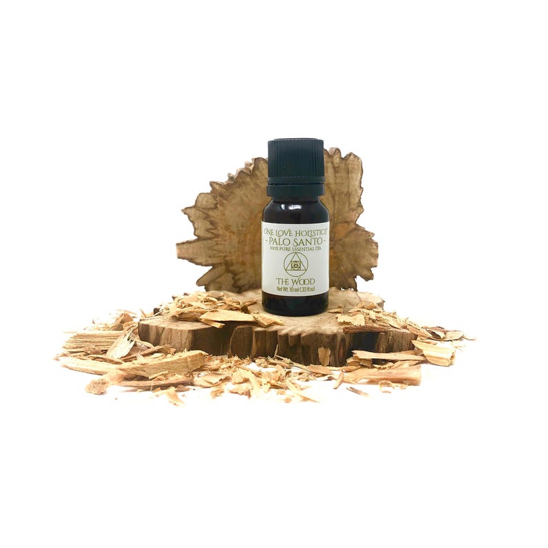 Op de afbeelding: Een klein bruin glazen flesje met een zwarte dop, gevuld met Palo Santo etherische olie. Het flesje staat op een stuk hout met houtkrullen eromheen. Het etiket vermeldt "One Love Holistic Palo Santo 100% Pure Essential Oil The Wood 10 ml (0.33 fl oz)"