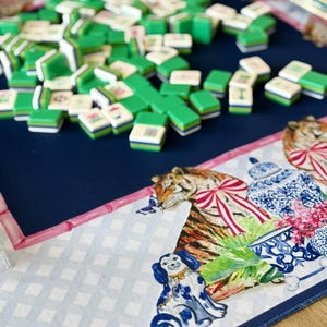 May include: A Mahjong set with green and white tiles scattered on a navy blue playing surface. The surface has a decorative border with a tiger, dog, and floral design. The tiles are stacked and ready for play.
