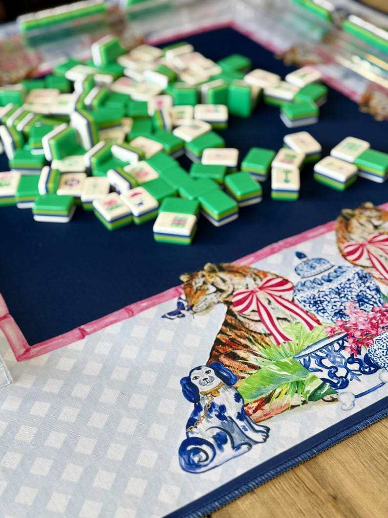 May include: A Mahjong set with green and white tiles scattered on a navy blue playing surface. The surface has a decorative border with a tiger, dog, and floral design. The tiles are stacked and ready for play.