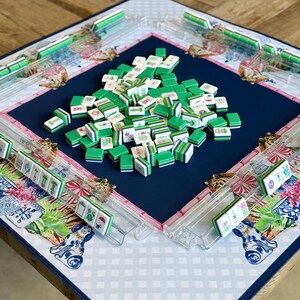 May include: A Mahjong game set with green and white tiles scattered on a navy blue playing surface. The board has clear plastic tile racks and is decorated with floral and animal patterns in pink, blue, and white.