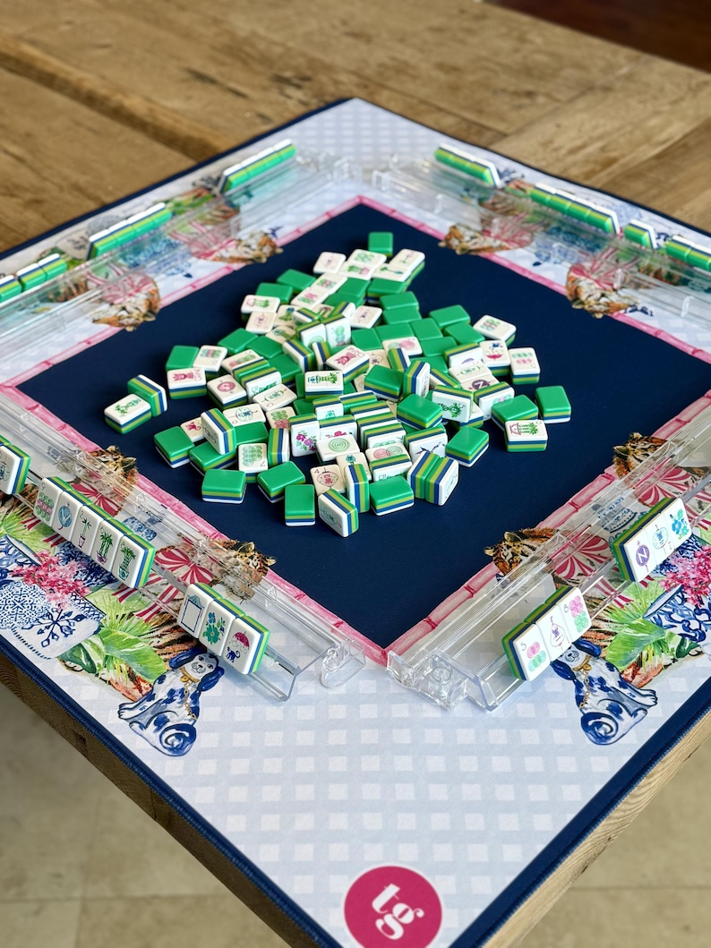 May include: A Mahjong game set with green and white tiles scattered on a navy blue playing surface. The board has clear plastic tile racks and is decorated with floral and animal patterns in pink, blue, and white.