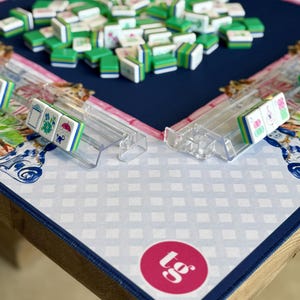 May include: A Mahjong set with green and white tiles scattered on a navy blue playing surface. Clear plastic tile racks are visible, holding a few tiles. The game is set on a table with a patterned mat and a pink circle with text.
