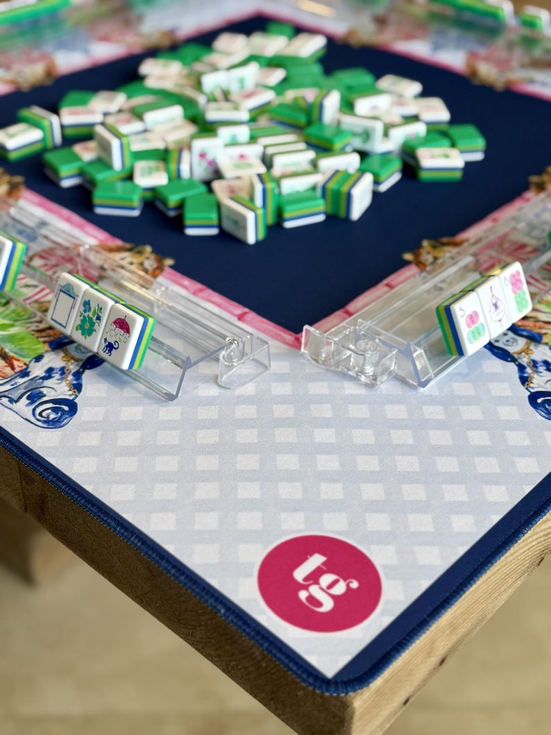 May include: A Mahjong set with green and white tiles scattered on a navy blue playing surface. Clear plastic tile racks are visible, holding a few tiles. The game is set on a table with a patterned mat and a pink circle with text.