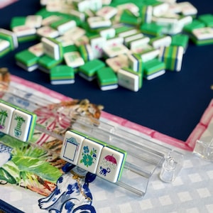 May include: A close-up of a Mahjong game in progress. The tiles are white with green and blue accents, featuring various symbols and designs. The game is set on a patterned surface with a blue background. Some tiles are in a clear plastic rack.