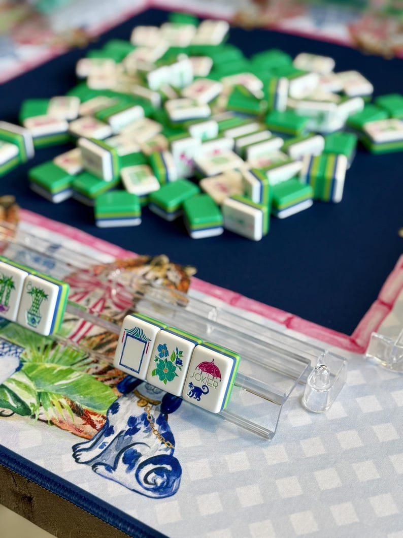 May include: A close-up of a Mahjong game in progress. The tiles are white with green and blue accents, featuring various symbols and designs. The game is set on a patterned surface with a blue background. Some tiles are in a clear plastic rack.