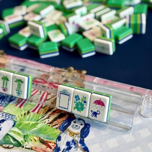 May include: A collection of green, white, and blue Mahjong tiles, some arranged in a row, others scattered. The tiles feature various symbols and illustrations, including flowers, a pagoda, and a monkey with an umbrella. A decorative mat with a blue and white dog is in the foreground.