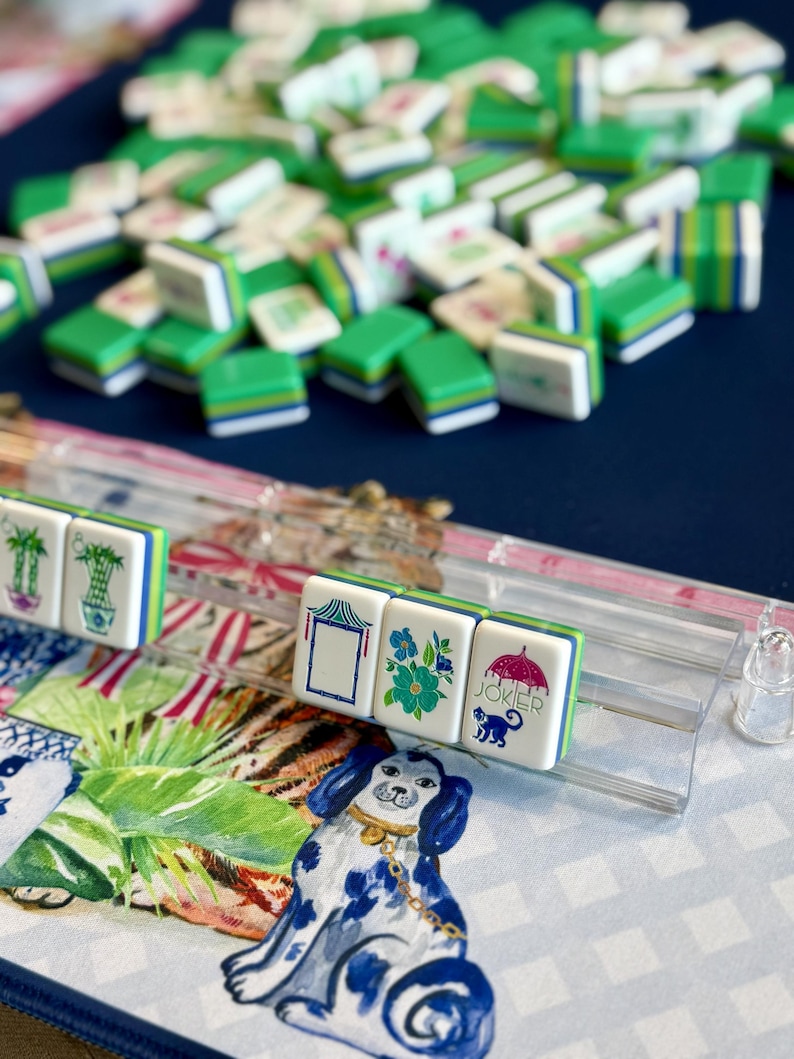 May include: A collection of green, white, and blue Mahjong tiles, some arranged in a row, others scattered. The tiles feature various symbols and illustrations, including flowers, a pagoda, and a monkey with an umbrella. A decorative mat with a blue and white dog is in the foreground.