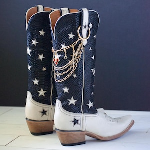 Könnte beinhalten: Ein Paar weiße Cowboy-Stiefel aus schwarzem Leder mit weißen Sternen. Die Stiefel haben goldene Ketten und Anhänger am oberen Rand.