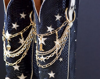 Può includere: Un paio di stivali da cowboy neri e bianchi con un motivo a stelle. Gli stivali hanno una texture a pelle di serpente e sono adornati con catene e ciondoli dorati. Gli stivali hanno un tacco e sono decorati con stelle bianche.
