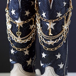 Puede incluir: Botas de vaquero con un estampado de piel de serpiente azul marino y estrellas blancas. Las botas tienen un pie de cuero blanco y están adornadas con cadenas y dijes dorados, incluyendo un corazón y una bota.