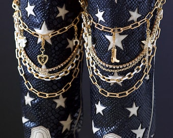 Può includere: Stivali da cowboy con un motivo a pelle di serpente blu navy e stelle bianche. Gli stivali hanno un piede in pelle bianca e sono adornati con catene e ciondoli dorati, tra cui un cuore e uno stivale.