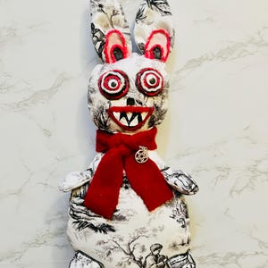 May include: A handmade plush rabbit with a black and white toile pattern. The rabbit has large, red-lined eyes, a red mouth with white teeth, and a red scarf. A silver pendant hangs from the scarf. The rabbit's ears are long and upright.