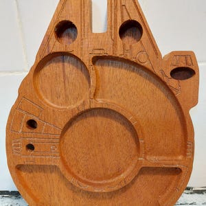 Puede incluir: Una bandeja de madera para servir con la forma del Halcón Milenario. La bandeja marrón presenta detalles tallados, incluyendo portavasos circulares y compartimentos. El diseño está inspirado en la icónica nave espacial de la franquicia Star Wars.