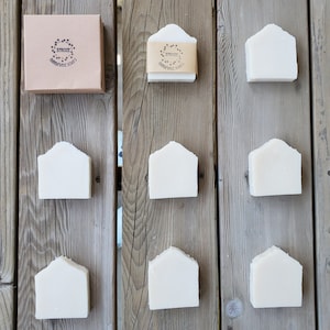 May include: Nine handmade soap bars in a house shape, arranged in three rows on a wooden surface. The soap bars are white and have a slightly textured surface. The top row also includes a brown cardboard box with the text "Strozzi Handmade Soaps" printed on it.