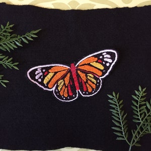 May include: Embroidered butterfly patch with orange, yellow, and red wings, outlined in black and white. The patch is set against a black background with green fern accents. A decorative item for clothing or accessories.
