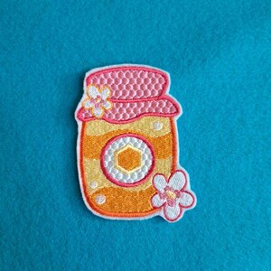 May include: Embroidered patch of a honey jar with a pink lid and a white and yellow flower. The jar is orange and yellow with a white and yellow hexagon in the center. The patch has an orange border and is on a blue background.