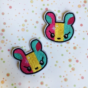 May include: Two embroidered iron-on patches featuring a cartoon bunny with pink, yellow, and teal colours. The bunny has a mischievous expression with black eyes and a small smile.