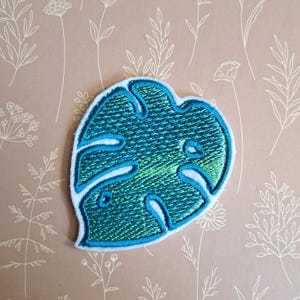 May include: A teal blue and green embroidered monstera leaf patch. The patch is outlined in white and has a textured, stitched appearance.