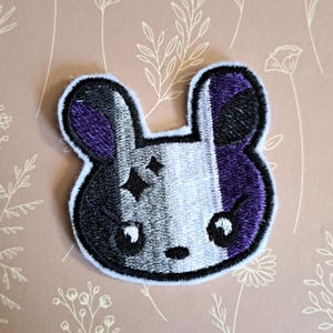 May include: A white, grey and purple embroidered bunny patch with a black outline. The bunny has a black nose and white eyes.