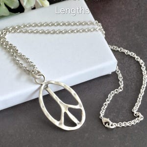 May include: A silver-coloured necklace featuring a peace sign pendant. The pendant is oval with the peace symbol. The chain has a clasp. The text "Choice of Chain Lengths" is visible. The necklace is a stylish accessory.