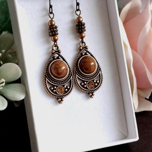 May include: A pair of bronze-coloured drop earrings. Each earring has a teardrop shape with a central brown stone, framed by a decorative rope-like border. Small amber-coloured crystals and beaded accents add detail.