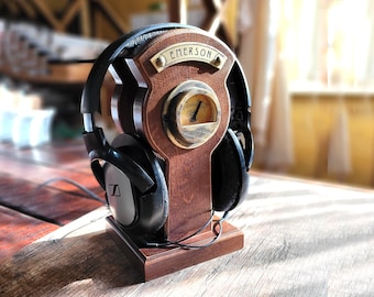 Puede incluir: Un soporte para auriculares de madera oscura con una placa de latón con el texto EMERSON y un indicador analógico circular centrado en la parte frontal. Unos auriculares negros descansan sobre el soporte, que tiene una base rectangular.