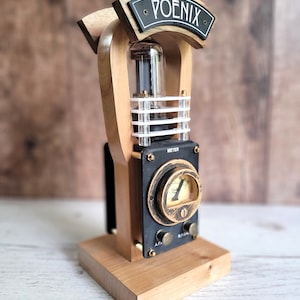 以下が含まれることがあります： 「POENIX」の文字が書かれた黒い看板が付いた、木と金属の手作りアート作品。 真空管、メーター、真鍮のアクセントがデザインに取り入れられています。 ベースは明るい色の木で、全体的な美学はスチームパンクです。