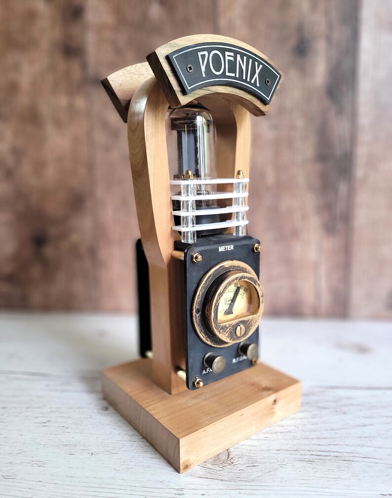 以下が含まれることがあります： 「POENIX」の文字が書かれた黒い看板が付いた、木と金属の手作りアート作品。 真空管、メーター、真鍮のアクセントがデザインに取り入れられています。 ベースは明るい色の木で、全体的な美学はスチームパンクです。