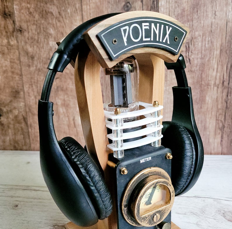 以下が含まれることがあります： 黒と金のメーター、透明なチューブ、そして「POENIX」の文字が書かれた黒い看板が付いた木製のヘッドホンスタンド。黒いヘッドホンがスタンドに置かれています。スタンドはヴィンテージ、スチームパンクの美学を持っています。
