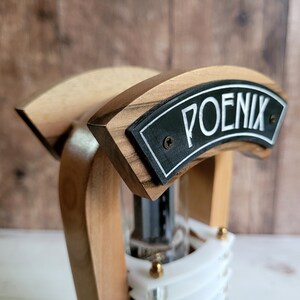 以下が含まれることがあります： 黒いプレートに白い文字で「POENIX」と書かれた木製のバータップハンドル。ハンドルは、明るい色と暗い色の木材を使った曲線的なデザインで、透明なガラス管が見えます。