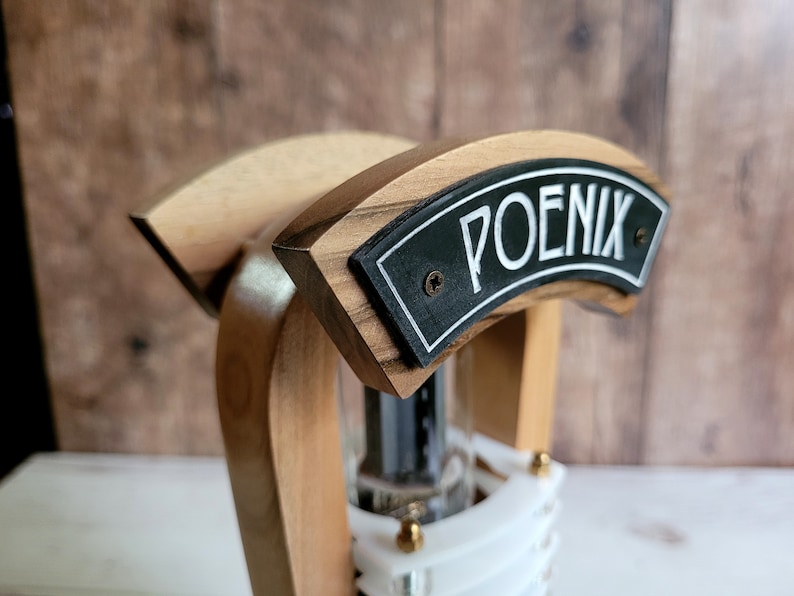 以下が含まれることがあります： 黒いプレートに白い文字で「POENIX」と書かれた木製のバータップハンドル。ハンドルは、明るい色と暗い色の木材を使った曲線的なデザインで、透明なガラス管が見えます。