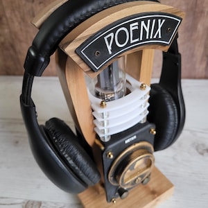 以下が含まれることがあります： 「POENIX」の文字が書かれた黒いプレートが付いた木製のヘッドホンスタンド。スタンドには黒と金のメーター、透明なチューブ、革製のイヤーカップが付いた黒いヘッドホンがあります。スタンドは明るい色の木材で作られています。