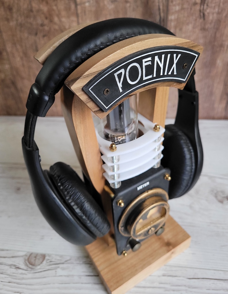 以下が含まれることがあります： 「POENIX」の文字が書かれた黒いプレートが付いた木製のヘッドホンスタンド。スタンドには黒と金のメーター、透明なチューブ、革製のイヤーカップが付いた黒いヘッドホンがあります。スタンドは明るい色の木材で作られています。